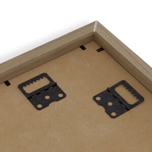 May include: Close-up of the back of a wooden picture frame. The frame has a light brown finish and features two black metal sawtooth hangers and several black staples. The backing is a light brown color.