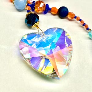 May include: A crystal heart-shaped prism with iridescent rainbow colors hangs from a beaded chain with blue, orange, and light blue beads.