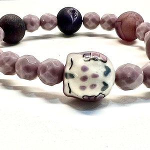 May include: A beaded bracelet featuring a central ceramic owl bead with purple and blue accents. The bracelet includes faceted lavender beads, dark purple and brown wooden beads. A unique handmade jewelry piece.