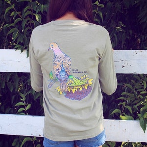 May include: A light green long-sleeved t-shirt featuring a graphic of a dove perched on a sunflower. The text "Ellie Mcginnis Co." is printed on the shirt.