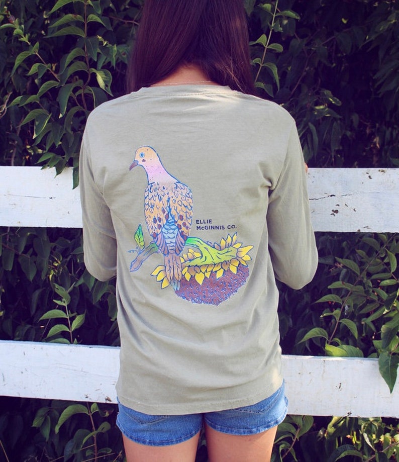 May include: A light green long-sleeved t-shirt featuring a graphic of a dove perched on a sunflower. The text "Ellie Mcginnis Co." is printed on the shirt.