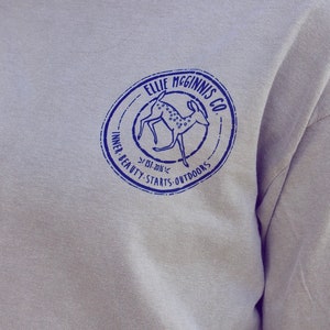 May include: A grey long-sleeved shirt with a blue circular logo featuring a deer and the text "Ellie McGinnis Co. Est. 2014 Inner Beauty Starts Outdoors".