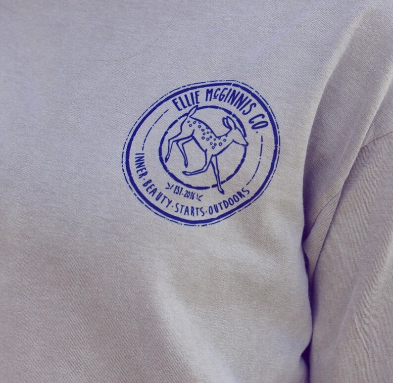 May include: A grey long-sleeved shirt with a blue circular logo featuring a deer and the text "Ellie McGinnis Co. Est. 2014 Inner Beauty Starts Outdoors".