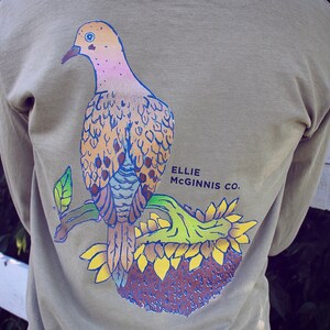 May include: A long-sleeved shirt in a muted green colour features a graphic of a dove perched on a sunflower. The dove is depicted in shades of pink, brown, and blue. The text "ELLIE MCGINNIS CO." is printed in black below the dove.