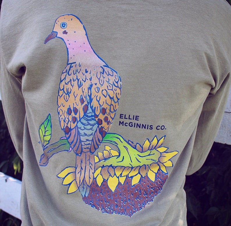 May include: A long-sleeved shirt in a muted green colour features a graphic of a dove perched on a sunflower. The dove is depicted in shades of pink, brown, and blue. The text "ELLIE MCGINNIS CO." is printed in black below the dove.