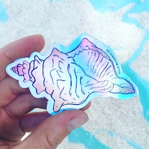 May include: A holographic sticker of a pink seashell with a blue outline. The sticker has the text "ELLIE MCGINNIS CO." printed on the right side.