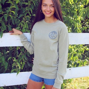 May include: A light green long-sleeved t-shirt with a blue circular logo on the left side. The logo features a bird with a compass and the text "The Journey".