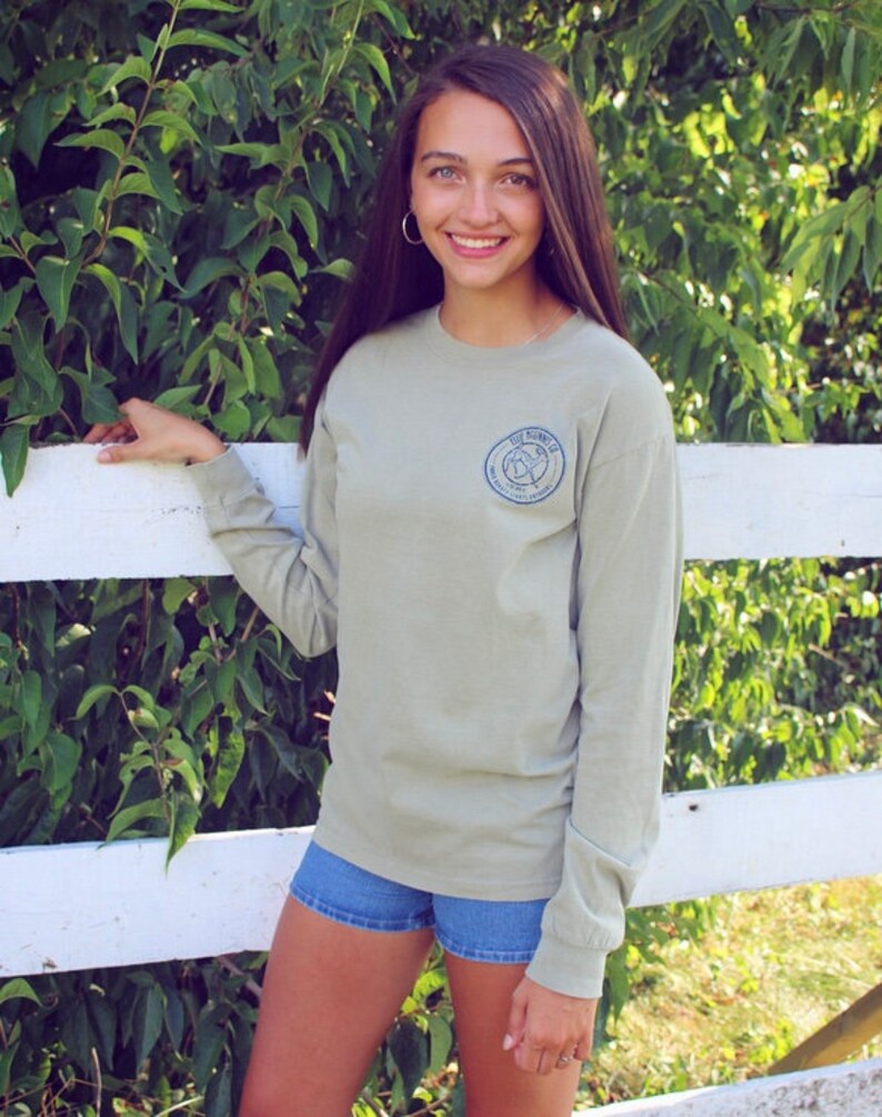 May include: A light green long-sleeved t-shirt with a blue circular logo on the left side. The logo features a bird with a compass and the text "The Journey".