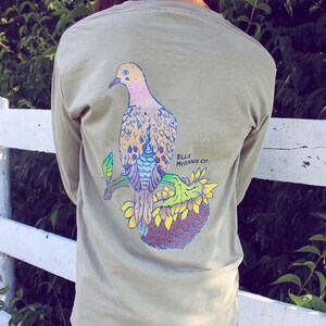 May include: A light green long-sleeved t-shirt featuring a graphic of a dove perched on a sunflower. The text "Ellie Mcginnis Co." is printed on the shirt.