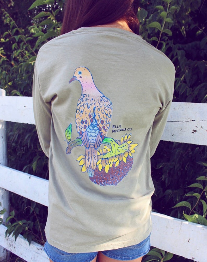 May include: A light green long-sleeved t-shirt featuring a graphic of a dove perched on a sunflower. The text "Ellie Mcginnis Co." is printed on the shirt.