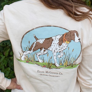 May include: Cream-colored long-sleeve shirt featuring a hunting dog illustration within an oval frame. The dog is brown and white, with a yellow collar, set against a light blue background. Text on the shirt reads "Ellie McGinnis Co. - Hunting Collection."