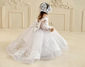 May include: A white tulle dress with lace detailing and a matching lace bonnet. The dress has a large bow at the back and long sleeves.
