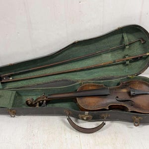 May include: An antique violin in an open, dark green-lined case. The violin is brown with a dark finish, and two bows are visible. The case is dark brown with a handle and metal clasps.