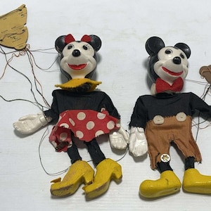 May include: Two vintage marionette puppets featuring Minnie and Mickey Mouse. Minnie wears a black top, red skirt with white polka dots, and yellow heels. Mickey wears a black top, brown pants with two buttons, and yellow shoes. Both have control bars.