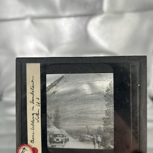 May include: A vintage black and white glass lantern slide featuring a landscape with a large sandstone rock formation, a parked car, and two figures on a road. A paper label on the left side contains handwritten text reading Cross bedding in Sandstone Utah U.S.