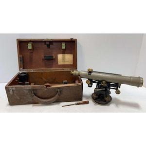 May include: A vintage surveying level instrument with a metallic gray telescope and black base, featuring brass adjustment knobs. It is accompanied by a wooden carrying case with a metal plaque and a small screwdriver with a wooden handle.