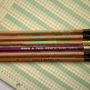 May include: Several pencils with erasers are laid out on a grid-lined surface. The pencils are mainly brown and purple, with text in green and gold. The erasers are in various colours, including green, pink, and white.