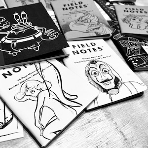 Puede incluir: Una colección de cuadernos ilustrados en blanco y negro con varios diseños, incluyendo un cangrejo, una persona con una máscara y un personaje de dibujos animados. Los cuadernos están etiquetados como "Field Notes" y "48-Page Memo Book".