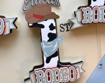 May include: A cake topper featuring a black and white cow print number one with a blue bandana, a brown cowboy hat, and a red and white banner that says "RODEO" and "1st". The topper is decorated with the name "Elliott's" in a brown script font.