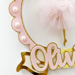 Op de afbeelding: Roze en gouden taarttopper met een ballerina-silhouet, een roze tule rok en de naam "Olivia" geschreven in roze met gouden glitteraccenten.