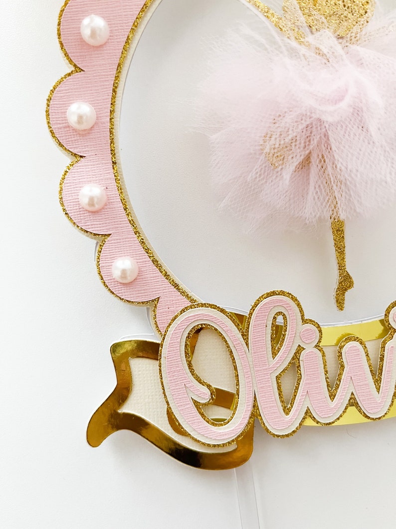 Op de afbeelding: Roze en gouden taarttopper met een ballerina-silhouet, een roze tule rok en de naam "Olivia" geschreven in roze met gouden glitteraccenten.
