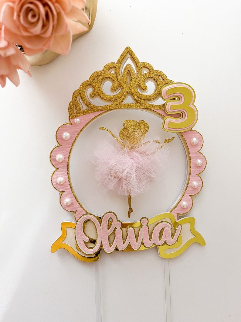 Op de afbeelding: Een roze en gouden ballerina taarttopper met een kroon, het nummer 3 en de naam Olivia. De ballerina draagt een roze tutu en heeft gouden glitteraccenten.
