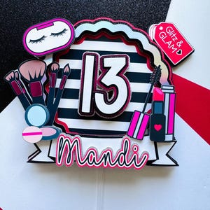 May include: A colorful cake topper with a makeup theme. The design includes a black and white striped background with the number 13, the name "Mandi", and makeup-related graphics like brushes, lipstick, and an eye shadow compact. A tag reads "Glitz & GLAM".