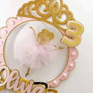 Op de afbeelding: Een roze en gouden taarttopper met een ballerina-silhouet, een kroon, het nummer 3 en de naam "Olivia".