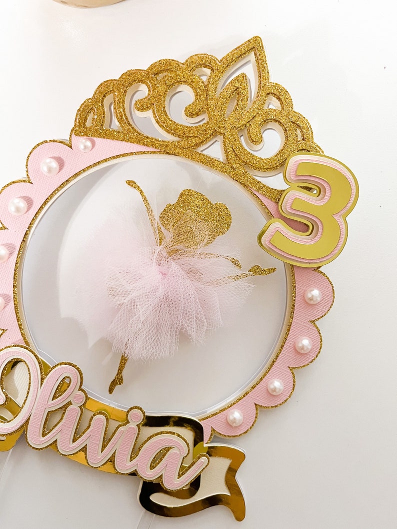 Op de afbeelding: Een roze en gouden taarttopper met een ballerina-silhouet, een kroon, het nummer 3 en de naam "Olivia".