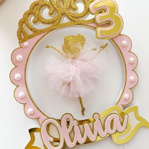 Op de afbeelding: Een roze en gouden taarttopper met een ballerina-silhouet, een kroon, het nummer 3 en de naam Olivia.