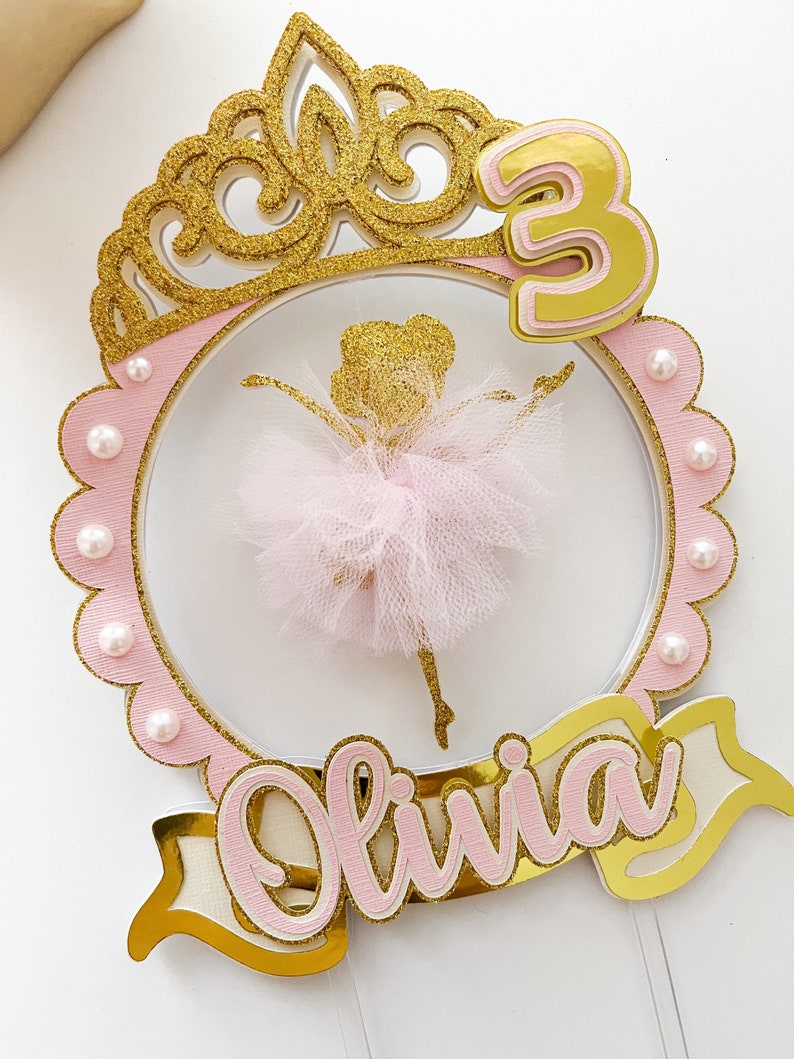 Op de afbeelding: Een roze en gouden taarttopper met een ballerina-silhouet, een kroon, het nummer 3 en de naam Olivia.