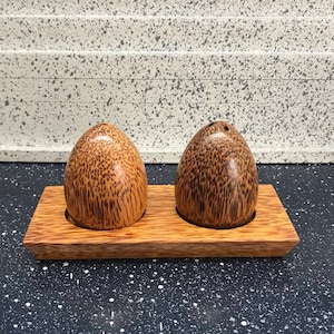 以下が含まれることがあります： 卵の形をした木製の塩と胡椒入れが2つ、木製のトレイの上に置かれています