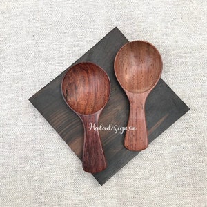 May include: Two wooden spoons with rounded bowls and short handles rest on a dark, square wooden board. The spoons are a rich, reddish-brown color, showcasing the natural wood grain. The board sits on a light-colored, textured surface.