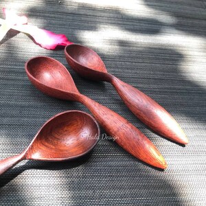 May include: Three wooden spoons with a reddish-brown color, made of a dark wood, are arranged on a gray woven surface. The spoons are all different sizes and have a rounded bowl shape.