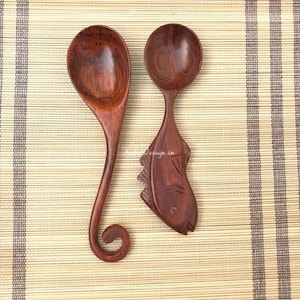 May include: Two wooden spoons, one with a curved handle and the other carved into the shape of a fish. The spoons are on a woven bamboo mat.
