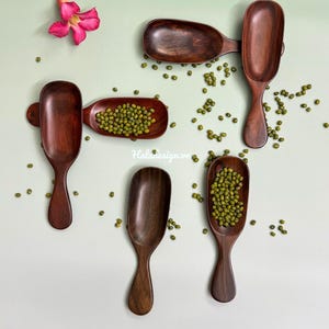 May include: A collection of handcrafted wooden scoops and serving dishes, some filled with green mung beans, arranged on a light green surface. A pink flower and scattered beans add color to the composition. The scoops vary in size and shape.