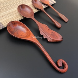 May include: Set of four wooden spoons, two with a simple rounded handle and two with a more elaborate carved handle. One spoon is carved in the shape of a fish. The spoons are a reddish brown colour.