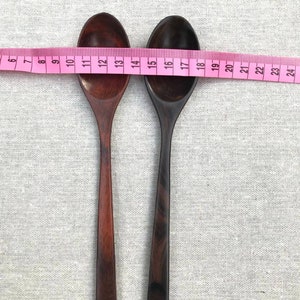 Op de afbeelding: Twee houten lepels, een donkerbruin en een roodbruin, liggen naast elkaar op een wit stofoppervlak. Een roze meetlint is over de lepels gelegd, met de nummers 7 tot en met 24 zichtbaar.
