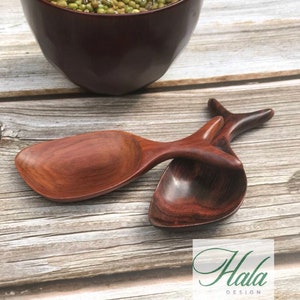 Può includere: Due cucchiai di legno a forma di pesce, uno marrone e uno marrone rossiccio, sono appoggiati su una superficie di legno. I cucchiai sono realizzati in legno scuro e hanno una finitura liscia. I cucchiai sono un modo unico e decorativo per servire il cibo.