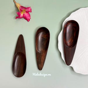 Könnte beinhalten: Drei dunkle Holzlöffel mit glatter Oberfläche werden auf einer hellgrünen Oberfläche zusammen mit einer rosa Blume und einem weißen Zierteller präsentiert. Die Löffel variieren leicht in Größe und Form und zeigen die natürliche Holzmaserung.
