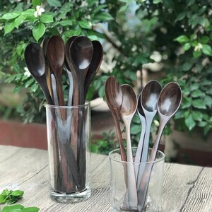 Op de afbeelding: Een set donkerbruine houten lepels in twee glazen potten. De lepels zijn verticaal in de potten gerangschikt, met de handvatten naar boven gericht. De potten staan op een houten oppervlak.