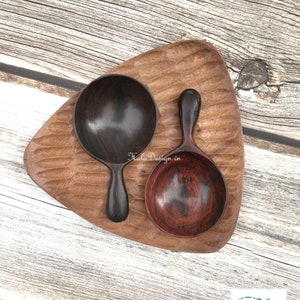 May include: Two wooden spoons, one dark brown and one reddish brown, are displayed on a triangular wooden tray. The spoons are handcrafted and have a unique, natural wood grain.