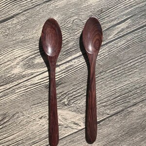 May include: Two wooden spoons with dark brown wood grain. The spoons are laying flat on a light gray wood surface.