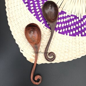 May include: Two wooden spoons with a unique swirl design at the handle. The spoons are brown and have a natural wood finish. The spoons are on a woven background with purple and white stripes.