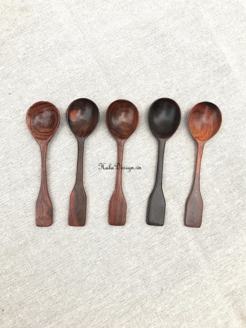 May include: Five wooden spoons with different wood grain patterns. The spoons are arranged in a row on a light grey fabric background.