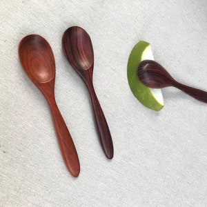May include: Three wooden spoons with dark brown and reddish brown wood grain. The spoons are on a white linen background.