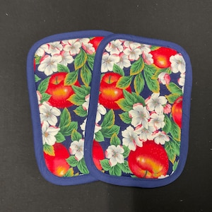 May include: Two rectangular pot holders with a vibrant apple and floral pattern. The design features red apples, white blossoms, and green leaves on a navy blue background, framed by a navy blue border. These kitchen accessories are designed to protect surfaces from heat.