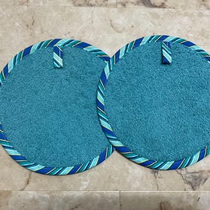 May include: Two round, teal-colored fabric pot holders with a textured surface. Each pot holder features a blue, green, and yellow diagonal striped fabric trim around the edge and a small matching fabric loop at the top for hanging purposes.