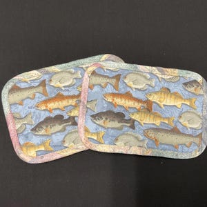 May include: Two rectangular pot holders with a light blue background featuring various fish illustrations. The edges are trimmed with a patterned fabric. The word "OREGON" is visible on the pot holders.
