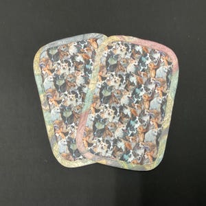 May include: Two dog-themed pot holders with a colorful pattern of various dog breeds. The pot holders have a rounded rectangular shape with a patterned border in shades of green, pink, and yellow.